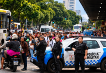 Imagem ilustrativa gerada por inteligência artificial de policiais militares, viatura e multidão em via urbana, representando o debate sobre segurança pública nas eleições de 2026